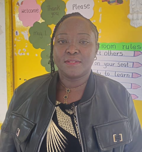 Woman in a gray leather jacket posing in front of a classroom bulletin board with colorful polite phrase decorations