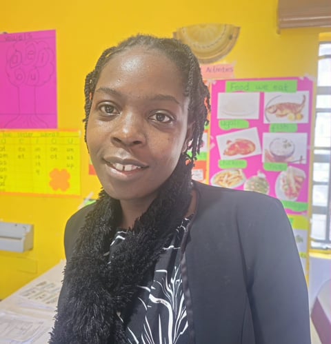 A woman smiling at the camera in a classroom with colorful educational posters and materials on yellow walls behind her