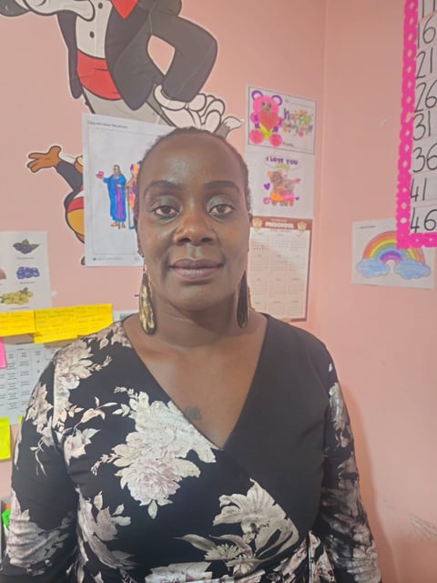 A woman wearing a black floral top and gold hoop earrings smiles at the camera in a classroom with pink walls and childrens artwork displayed.