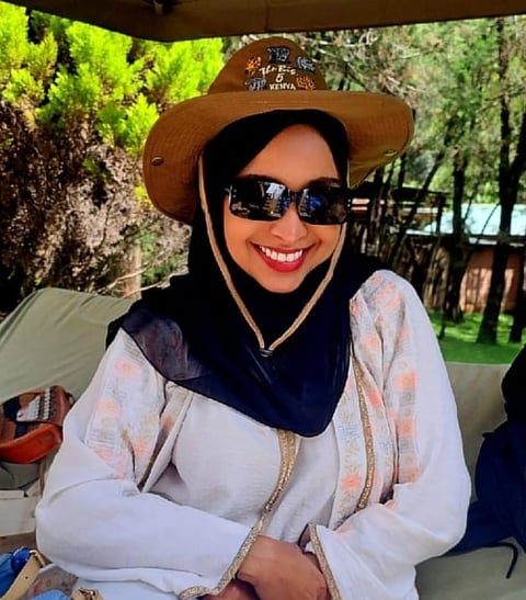 Woman wearing brown cowboy hat, black sunglasses, white shirt, and black hijab smiling at outdoor event with green trees in background